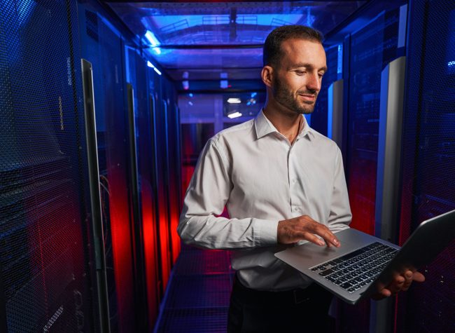 Caucasian programmer is looking to the screen of laptop while standing near the server rack